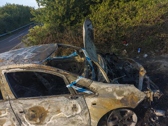A burnt out and weathered car just off a road has police tape wrapped around it, bonnet open and caved in, with space where an engine should be. Its wheels are just hubcaps.