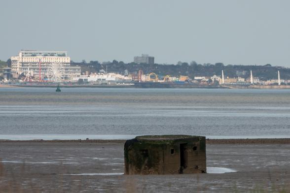 A weathered concrete pillbox sits on mudflats. Across a wide river there's a seaside resort, with funfair and tall hotels.