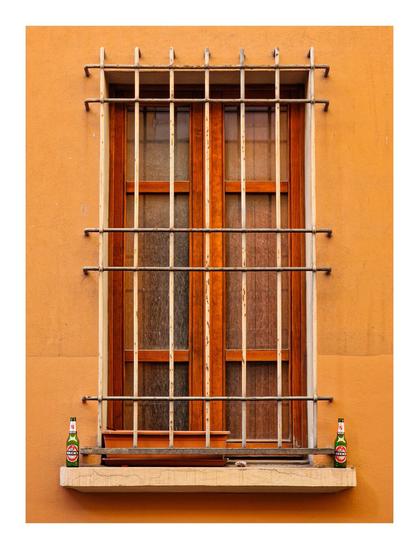 The photo is of a closed, wooden-framed window against an orange wall. The window is protected by vertical and horizontal metal bars. On the window sill, there are two identical green glass beer bottles (Beck's) placed symmetrically - one on each end. Inside the metal bars between the bottles is a empty rectangular flower pot.
