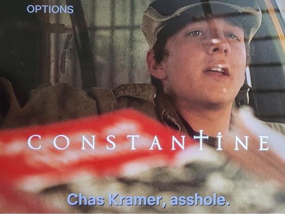 CONSTANTINE
Shows Chas Kramer in a flat top hat trying to look tough as he looks at himself in the rear view mirror of a car. He says "Chas Kramer, asshole."