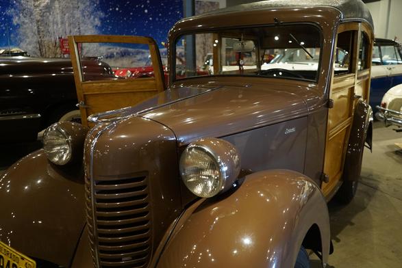 The image features a vintage brown car, likely from the 1930s or 1940s, prominently displayed in what appears to be a museum or exhibition setting. The car has a classic design with a rounded front, large circular headlights, and a prominent vertical grille. The body is painted in a glossy brown finish, and the car has wood paneling on the sides, which is characteristic of station wagons from that era. The driver's side door is open, revealing a glimpse of the interior, which includes a wooden dashboard and a steering wheel. The car's license plate is visible, reading "COLORADO 940-40." The background shows other vintage cars, suggesting a collection or exhibition. The lighting is bright, highlighting the car's polished surface and the surrounding environment.

Provided by @altbot, generated privately and locally using Ovis2-8B