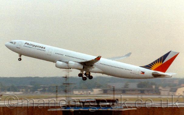 Slightly grainy side view of a white, 4 engined jet airliner flying from right to left at a very low altitude, with undercarriage lowered, flaps extended from the rear of the wings, and the nose raised sharply, suggesting it has just taken off.
The planes body is all white, with black "Philippines" titles on the upper forward fuselage, and blue, white, and red triangles on the tail overlaid with a yellow sun-burst.
A similar logo appears on the up-turned wing-tips.
The top of a brown building stretches across the bottom of the frame, with cream-coloured buildings in the distance, with trees beyond that, under grim grey sky.