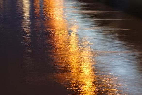 cCest une photo des reflets du soleil couchant dans un fleuve. L'image est très colorée allant d'un triangle noir  à la base élargie en bas à  gauche puis  passant à l'orange très vif avec un point focal jaune au centre de l'image. Une bande d'eau bleutée traverse en biais de la moitié du haut à l'angle droit du bas, avant de laisser la place à un triangle d'ombre  qui court le long du bord droit. Le contraste ombre/lumière et couleur froide/couleur chaude crée le dynamisme de la photo.