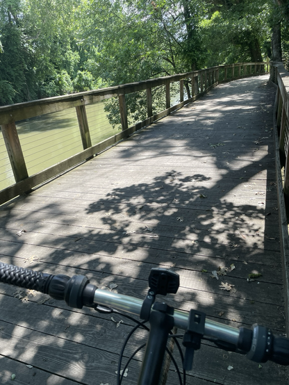 Pic overlooking bicycle handlebars on boardwalk next to small river, on a sunny day with tree shadows on boardwalk