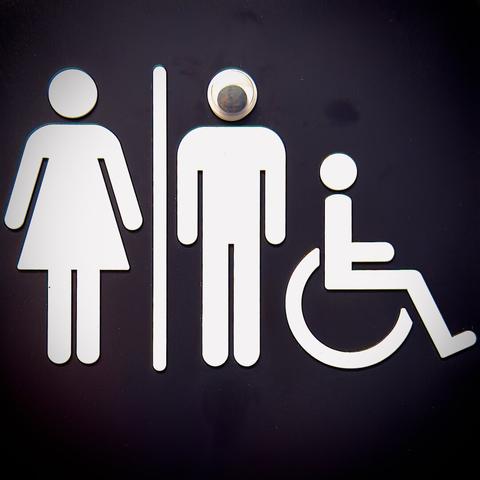A black bathroom placard with stick figures for a woman (dress), a man (no dress), and a person in a wheelchair—all displayed in white—has been tampered with a single googly eye stuck over the man's entire round head.