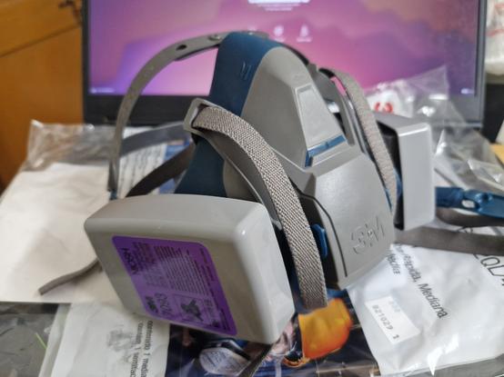 A blue-and-grey 3M 6502QL half-mask reusable elastomeric respirator, with attached 3M P100 7093 particulate cartridges, placed upright on a laptop keyboard.