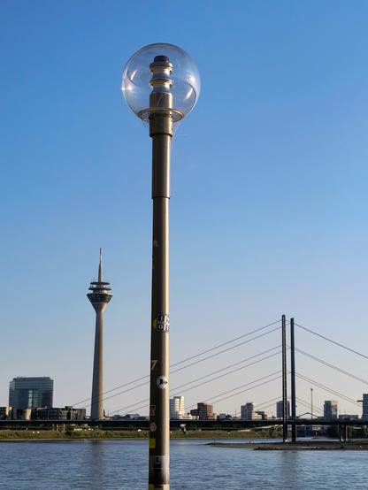 In der Mitte eine Kugel-Latere.
Im Hintergrund die Düsseldorfer Skyline mit blauem Himmel. Links der Fernsehturm, rechts zwei Brückenpfeiler. Unten Rheznwasser.