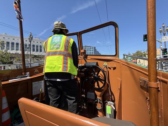 An operator in a neon yellow safety vest drives a vintage vehicle, with a green traffic light visible in the background. The interior is wooden and orange, showcasing a steering wheel and controls. The scene is set against a backdrop of buildings and clear blue sky
