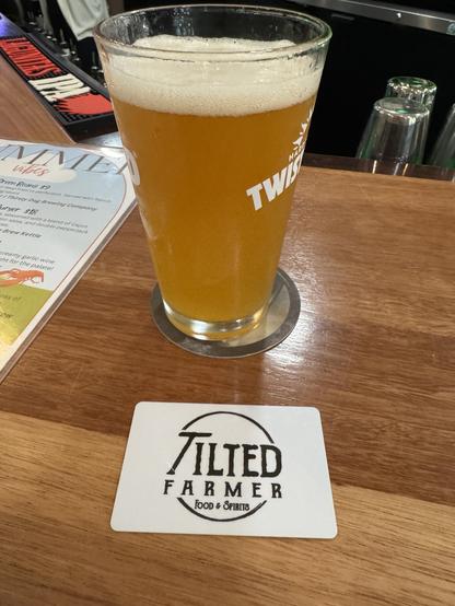 A glass of light-colored beer sits on a wooden bar countertop next to a card with the text "TILTED FARMER Food & Spirits." A menu is partially visible in the background.