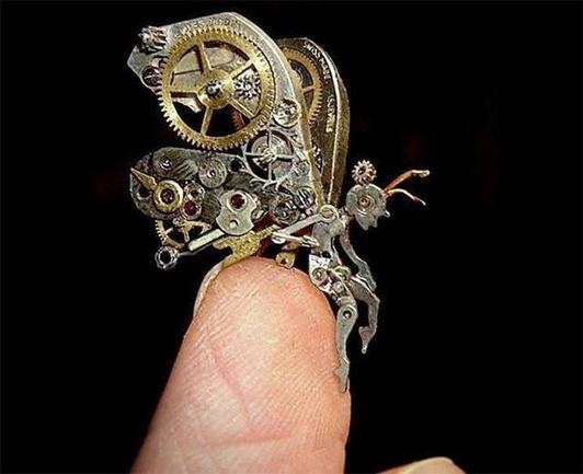 Close up photograph of a tiny sculpture resting on a white person''s fingertip, the artwork is in the shape of a sitting figure facing right with huge wings behind it, all made from tiny cogs and other very small watch parts, all against a black background