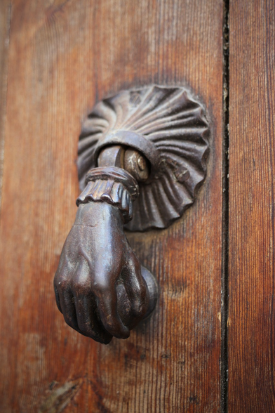A bronze doorknob shaped like a woman’s hand in a ruffled sleeve against a wooden door.
