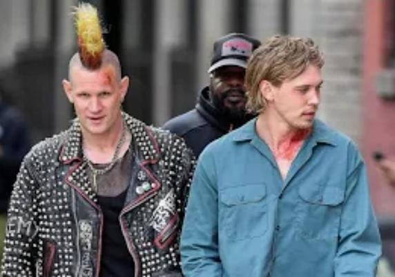 Matt Smith in a yellow 80s punk Mohawk and black leather studded motorcycle jacket. He's looking at the camera. Beside him is Austin Butler in a sky blue denim shirt and behind them is an unidentified gentleman in a dark baseball cap.