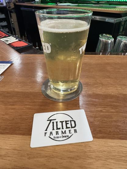 A glass of beer sits on a wooden bar counter next to a coaster featuring the logo of "Tilted Farmer Food & Spirits."