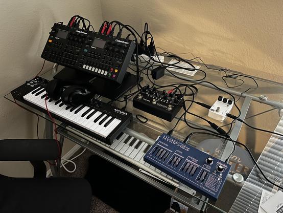 A bunch of music equipment (a keyboard, two grooveboxes, two effects pedals, and a small analog synth) on a desk with a tangle of too many cables hooking them all together