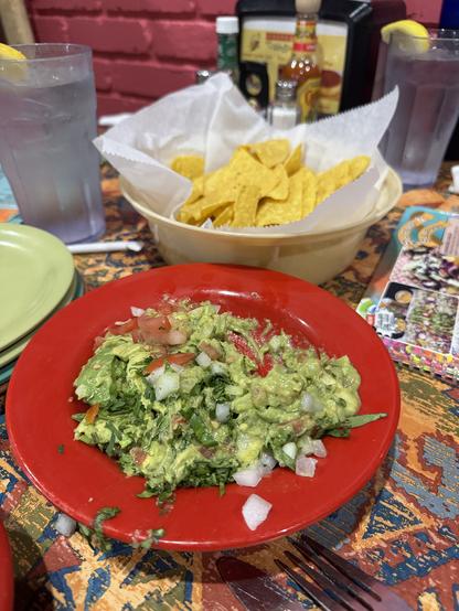 The image depicts a dining table setting with a focus on a red plate of guacamole in the foreground. The guacamole is chunky, with visible pieces of avocado, tomatoes, and onions. A fork is partially visible on the right side of the plate. Behind the guacamole, there is a beige basket filled with yellow tortilla chips lined with white paper. To the left, there are two glasses of water with lemon slices, and a stack of green plates. In the background, a red brick wall is visible, along with a condiment dispenser and a bottle of hot sauce. The tablecloth has a colorful, patterned design.

Provided by @altbot, generated privately and locally using Ovis2-8B

🌱 Energy used: 0.170 Wh