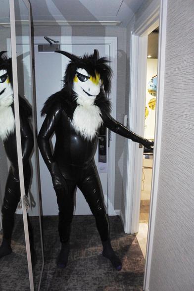 Fursuit photo - impromptu shot of a fluffy dragon wearing a polished black latex catsuit. He is standing in the hotel room hallway and is mostly lit by the camera flash.