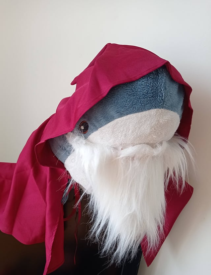 A blue and white IKEA Blahaj shark plushie wearing a red hooded robe and a long white beard, dressed up like a wizard or sage against a white background.