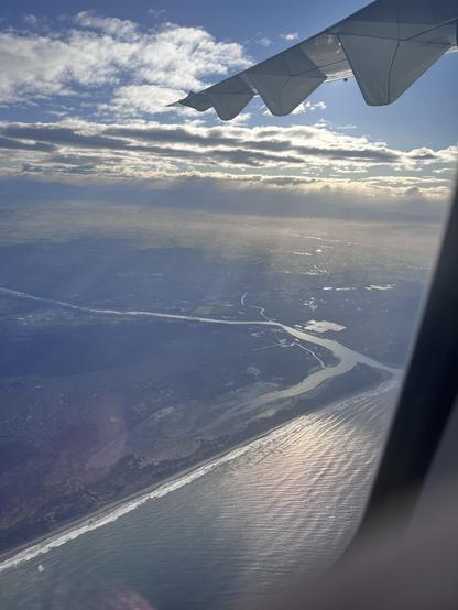 view out aircraft window of coastal scene with waves and rivers and clouds and a section of the wing - the sun is out of picture but very apparent in the way the haze lights up