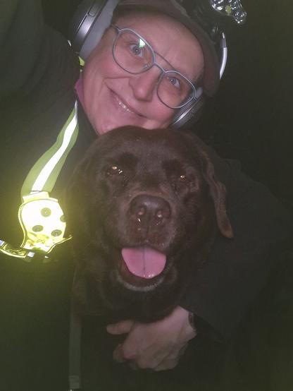 Night time selfie of a person and a chocolate brown Labrador with greying snout. The person is wearing a black jacket, yellow safety harness, red cap, silver headphones and blue-rimmed glasses. On their cap rests a headstrap torch in off position. Their free hand is around the dog's shoulders. Both are facing the camera. The dog's mouth is slightly open, showing off some blep. The person's head is resting on the dog's head. They're smiling, grateful for their time spent together.