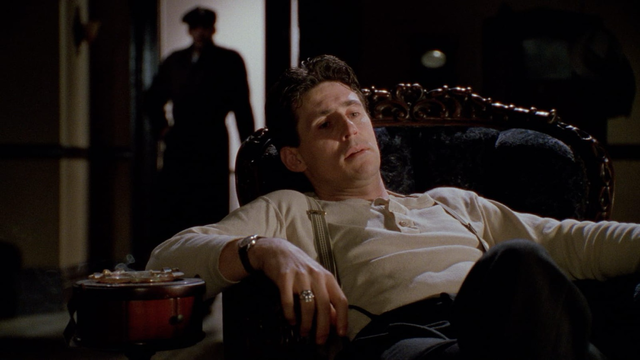 Gabriel Byrne sits pensively and non-ergonomically in chair while a menacing figure lurks in the background