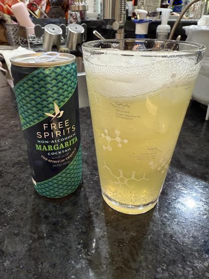 Photo with an empty can of Free Spirits non-alcoholic margarita cocktail on the left. Can also reads “the spirit of tequila.” On the right, a glass with ice and a yellow liquid, the non alcoholic margarita. The glass has images of molecules on it representing chemicals in beer