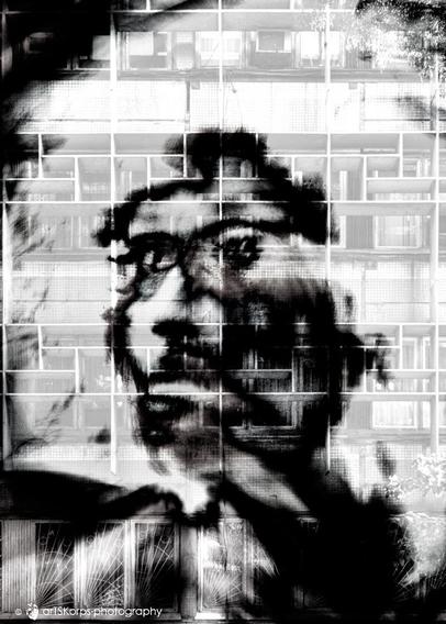 Black and white photo, portrait format, multiple exposure, self-portrait (headshot, motion blur, high-contrast). Against a light, textured background (the facade of an apartment block consisting of numerous windows), the silhouette of a face looking sideways, barely recognizable as such. The eyes appear to be sadly directed into the distance. The overall impression is one of hopelessness, despair, but also of holding oneself captive with one's own feelings.