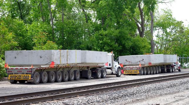 Two trucks with long flatbed trailers and "oversized load" banners carry four rectangular precast-concrete tunnel sections, perhaps 4 feet each. They wait alongside the Amtrak rails for crane crews to be ready to lift them off the trailers and set them near the tunnel site, for a construction blitz planned to begin Tuesday June 3rd.