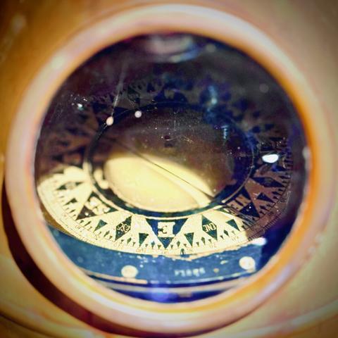 A large nautical compass is viewed through the circular port of its brass domed housing under dim tungsten lights.