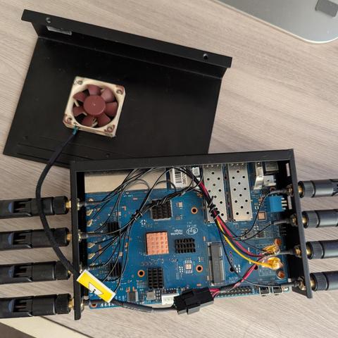 Una placa de circuito azul con numerosos componentes electrónicos está montada en una carcasa negra. La placa tiene varios conectores, cables y chips visibles. Se observa un pequeño ventilador cuadrado de color beige conectado a la carcasa. Cuatro antenas negras están conectadas a la parte posterior de la carcasa. La carcasa está parcialmente abierta, mostrando el interior. Una parte de la carcasa superior se encuentra separada, mostrando una superficie negra lisa.