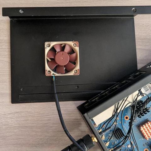 Una placa negra rectangular con un pequeño ventilador cuadrado de color beige y marrón está conectado a ella por medio de un cable.  Se ve parte de un dispositivo electrónico de color azul oscuro con numerosos cables y componentes en la parte inferior derecha de la imagen. El dispositivo parece ser una tarjeta de circuito.  La placa negra y el dispositivo electrónico están sobre una superficie de madera clara.