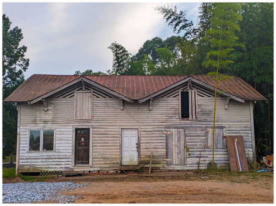 AI-assisted alt text:

A weathered, two-story wooden building (c. 1910) with peeling white paint and a rusty metal roof stands amid greenery. Several doors and windows are mismatched or boarded up. The ground in front is bare dirt with some gravel. Tall bamboo grows to the right side of the house.