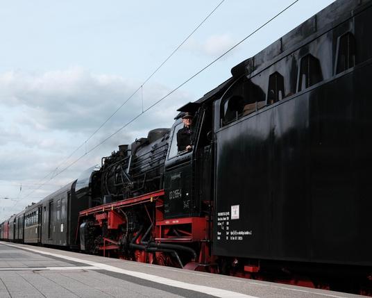 Eine Dampflokomotive mit der Nummer 03 2155-4 zieht einen Zug mit alten Personenwagen. Das Foto ist vom Bahnsteig aus niedriger Position und in schrägem Winkel aufgenommen. Die Lokomotive fährt mit dem Tender voran – der Kessel befindet sich hinten. Der Lokführer schaut aus dem Fenster nach vorn, am Betrachter vorbei. 

A steam locomotive with the number 03 2155-4 pulls a train with old passenger carriages. The photo was taken from the platform from a low position and at an oblique angle. The locomotive is running with the tender in front - the boiler is at the rear. The driver looks out of the window to the front, past the observer.