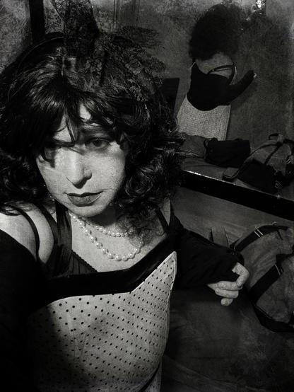 My photo is in black and white. I am wearing a Polka Dot dress. A small bed is behind me as well as a mirror. I am wearing a black wig looking at my camera.