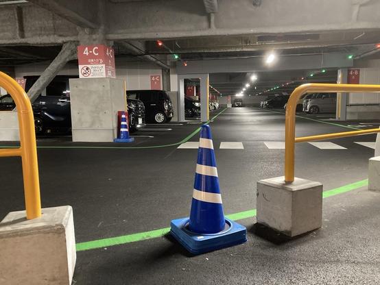 An underground parking area with designated spots, marked with green lines. A blue and white traffic cone is positioned in the foreground, and several parked vehicles are visible. Light fixtures illuminate the space, and signs indicate parking sections labeled "4-C".