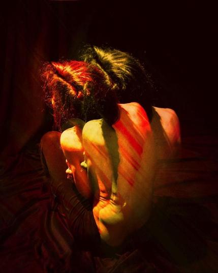 Color photograph, portrait format, multiple exposure. Self-portrait (torso viewed from behind) in red and orange tones against a dark background. The bent back of a person with long braided hair, legs bent while sitting, head tilted, additionally exposed with a shifted, identical photograph, so that the person is partially duplicated. Overall impression of splitness, seeking protection, self-protective posture, introverted. Fragile strength.