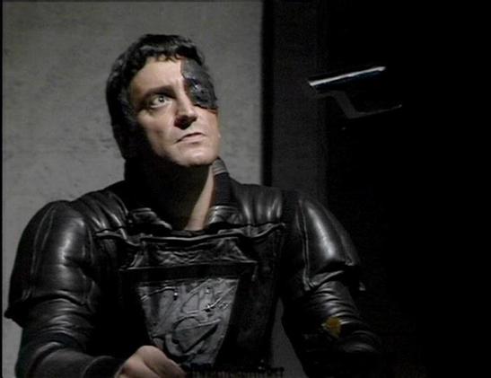 Claude Sonnet 4.0 describes the image as: "I can see this appears to be from a science fiction television production, showing someone in dark, futuristic military-style armor or uniform. The costume design has a distinctive sci-fi aesthetic with metallic elements and structured plating typical of space opera productions from that era. The lighting and set design create a dramatic, somewhat ominous atmosphere with dark tones and institutional-looking backgrounds. The overall production values and costume design suggest this is from a British science fiction series, likely from the late 1970s or early 1980s based on the visual style and filming techniques used."

Blakes7Bot, version 3.9.30.
