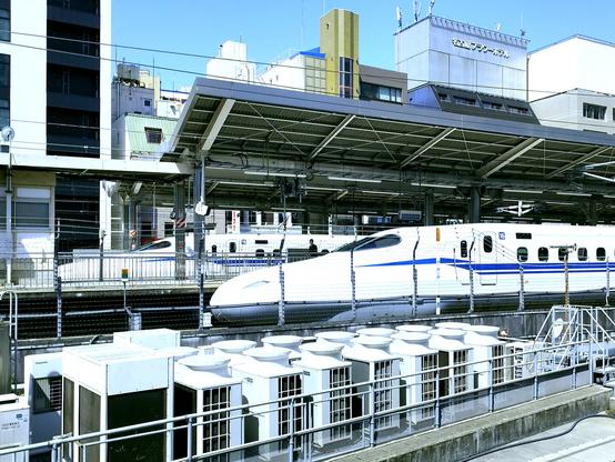 Among various items like A/C units and power lines as well as a cityscape background the picture shows two bullet trains calling at the station.