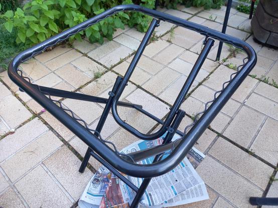 Frisch lackiertes Klapptischgestell auf einem Zeitungsblatt stehend. Darunter sieht man hellbraune Terrassenfliesen.
