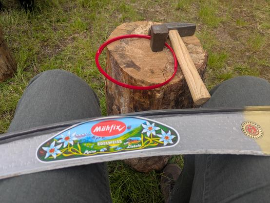 Ein Sensenblatt auf den Knien der fotografierenden Person, davor der Dengelamboss mit dem Dengelhammer