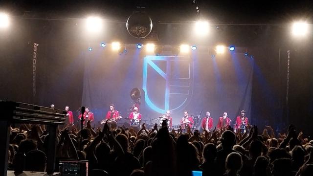 Musiker in roten Jacken eines Spielmannszugs auf einer Bühne. Das Bandlogo in blau. Im Vordergrund das Publikum, mit vielen nach oben gestreckten Armen.