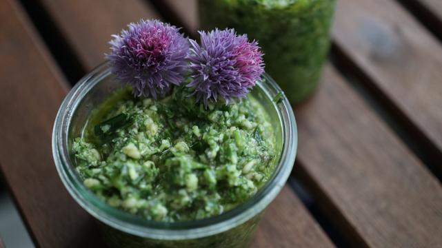 A jar with green pesto. On one side, two chive blossoms contrast the green with their hues of purple and pink.

---

Ein Glas grünes Pesto. Am Rand zwei Schnittlauchblüten, die mit ihrem lila und pink Kontrast zum Grün bieten.