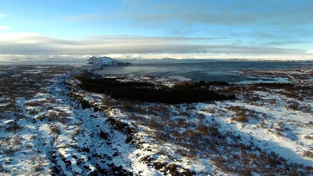A colour photo taken by a drone above a rugged landscape covered in snow with a lake in the distance. Dark rock is showing through the ice, and a deep cleft is running from bottom left to centre left, accentuated by shadows. The sky is cloudy but blue.