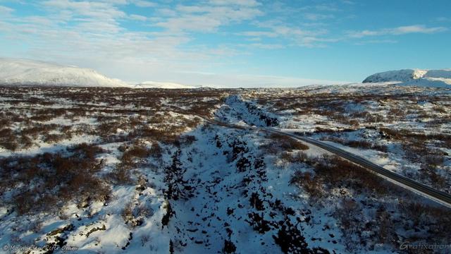 Another colour photo from a drone, showing the same snowy rift running from bottom centre and almost to the horizon. A road is running across from bottom right to centre left, dipping slightly as it crosses the chasm. There are snow-capped mountains on the horizon.