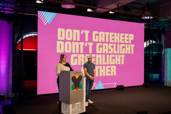 Zwei Personen stehen auf einer Bühne vor einer großen, pinken Leinwand mit der Botschaft: „DON’T GATEKEEP, DON’T GASLIGHT, GREENLIGHT GATHER.