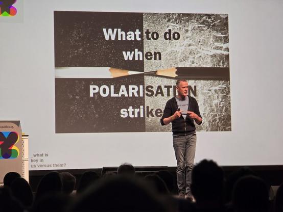 Ein Mann spricht auf einer Bühne vor einer Leinwand mit der Folie „What to do when POLARISATION strikes“ und dem Bild zweier zerbrochener Bleistifte.
