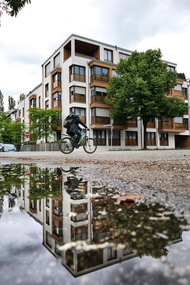 Das Bild zeigt ein modernes Wohngebäude mit mehreren Stockwerken und Balkonen. Die Fassade des Gebäudes ist überwiegend weiß, mit braunen Akzenten um die Fenster und Balkone. Eine Person fährt auf einem Fahrrad vorbei, trägt einen Rucksack und ist in Bewegung. Im Vordergrund befindet sich eine Pfütze, die eine beeindruckend klare Spiegelung des Gebäudes und der Fahrradfahrerin zeigt.