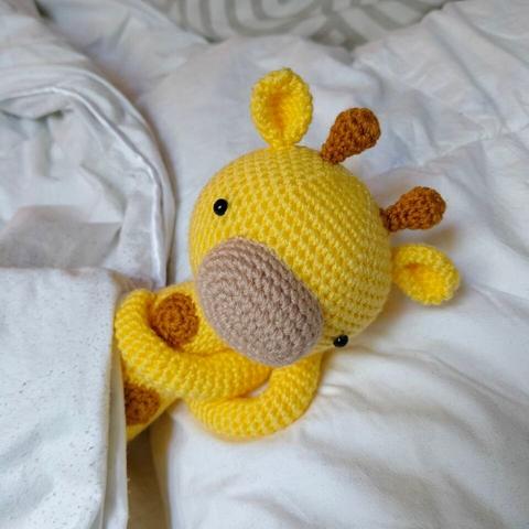 A picture of a crocheted amigurumi giraffe. It's lying down in a bed, under a blanket, only its upper part visible. Its arms are under its head.