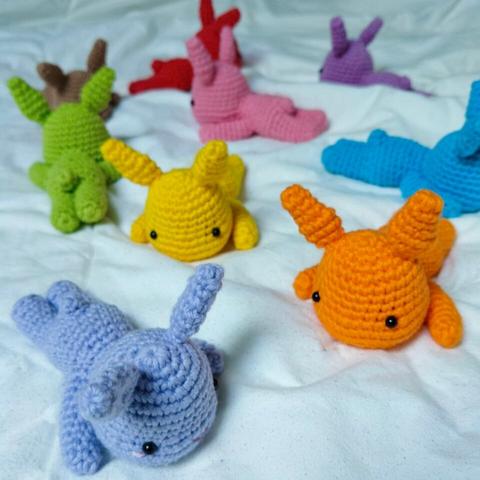 A picture of a variety of crocheted amigurumi bunnies in rainbow colors, all lying down on a white blanket on their bellies.