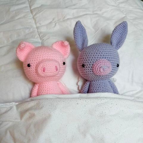 A picture of a crocheted pig and rabbit. They're in a bed, under a blanket, next to each other. They're only visible from the shoulders up.