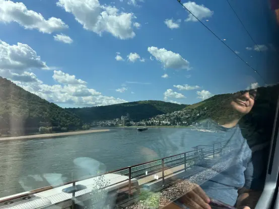 view down another Rhine curve, into a town with an old castle. My travel companion, @moof@cupoftea.social, is reflected in the window